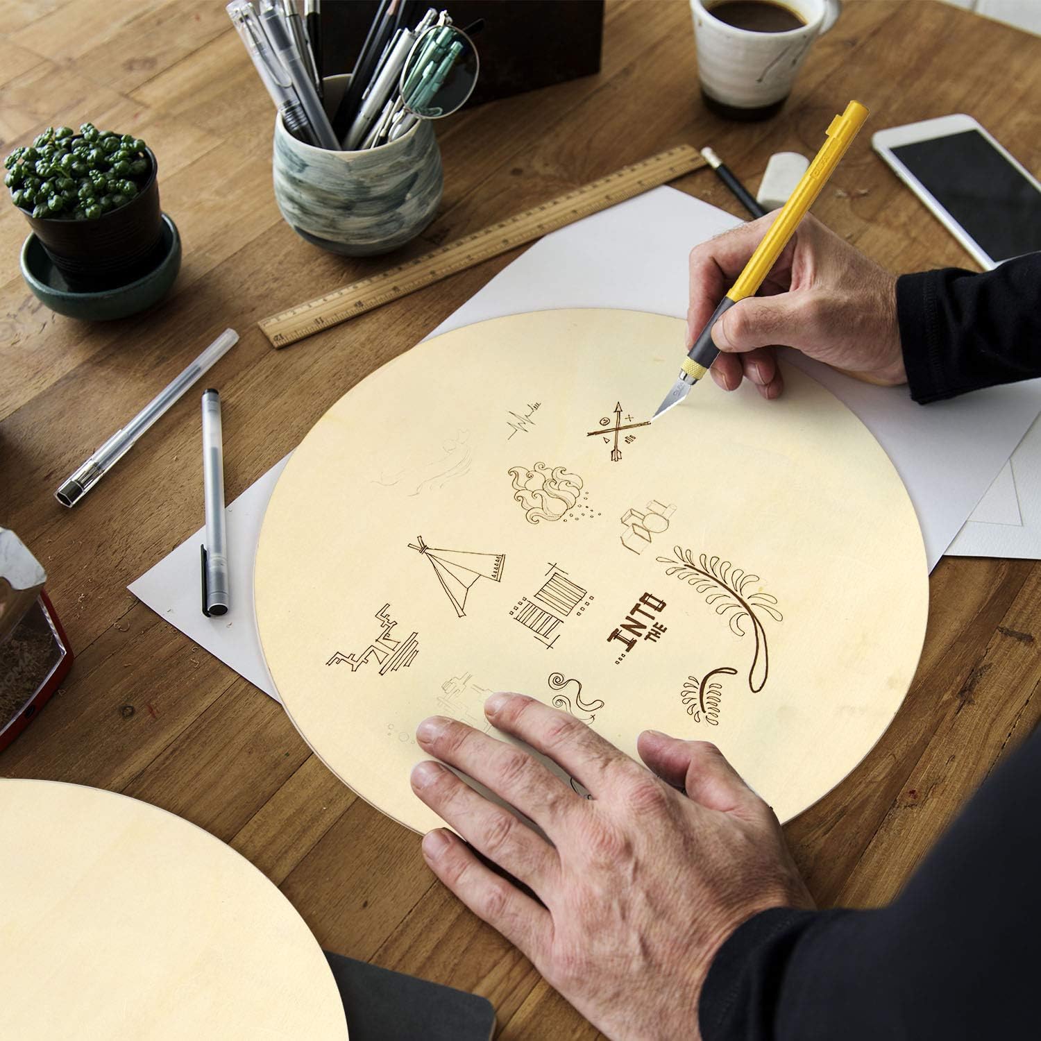 Person working on a wooden board with designs, using a pen on a desk with stationery items.