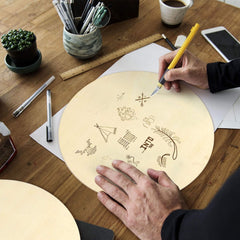 Person working on a wooden board with designs, using a pen on a desk with stationery items.