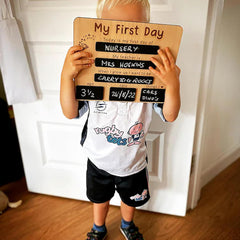 Child holding a 'My First Day' sign in front of their face, standing in a room.