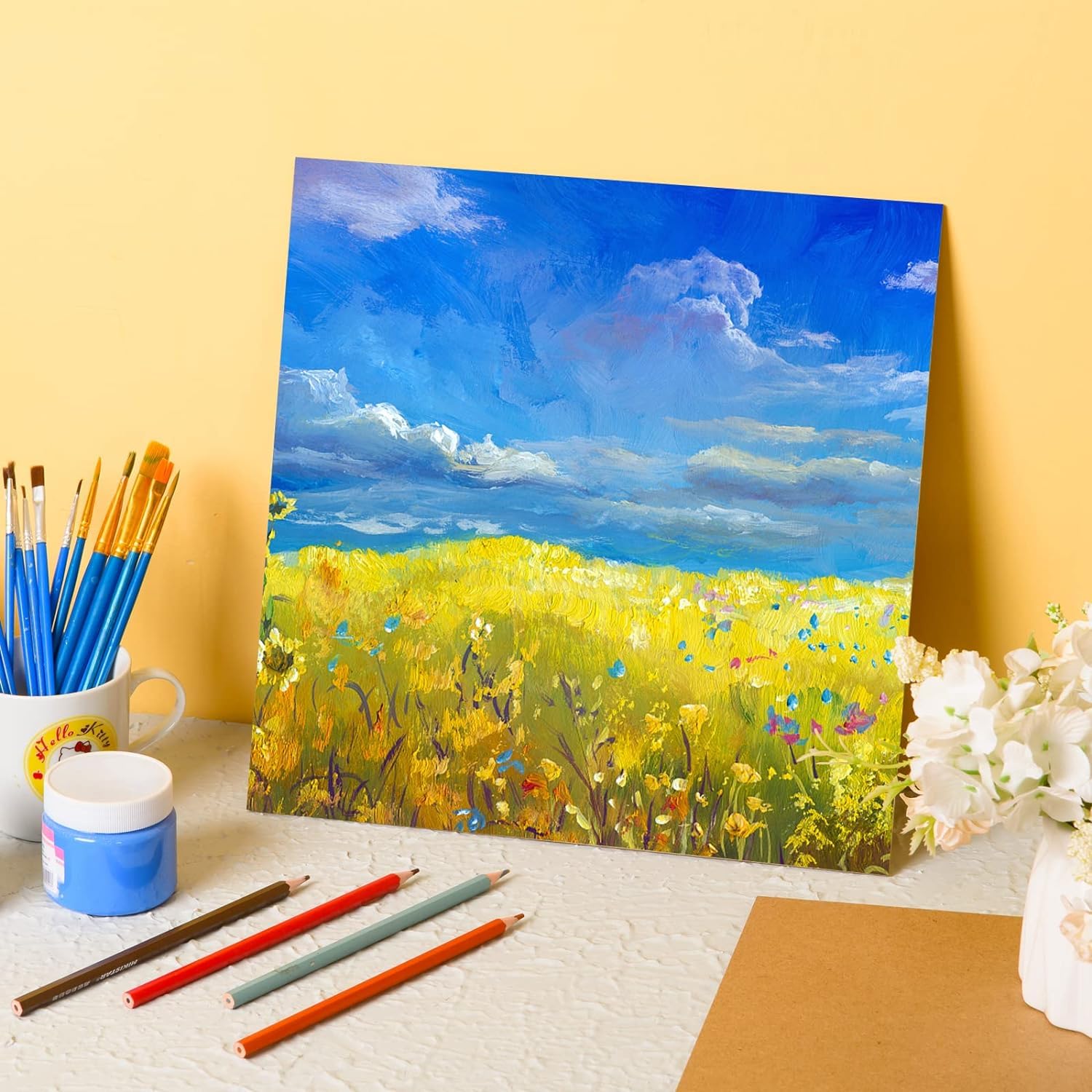 Painting of a field with flowers on a yellow background, surrounded by art supplies.