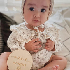 Baby in a floral outfit holding a 'nine months' milestone card