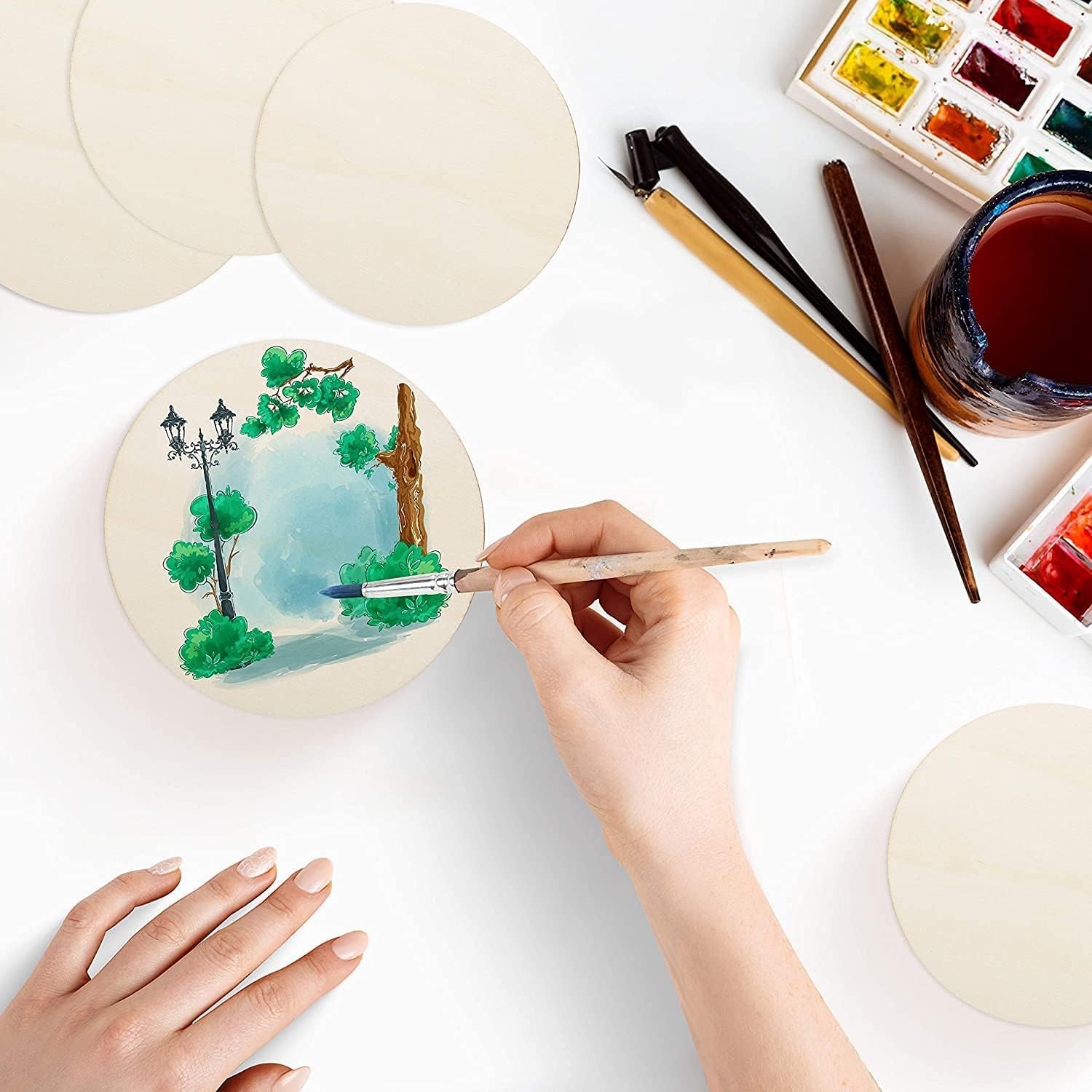 Person painting a small round canvas with trees and a sky, surrounded by art supplies on a white surface.