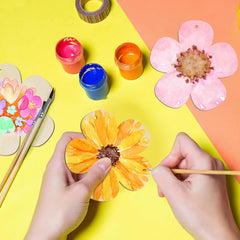 Person painting a flower-shaped craft on a colorful background with paint containers and brushes.