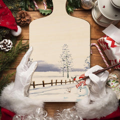 Person wearing white gloves holding a snowy landscape painting with Christmas decorations around.