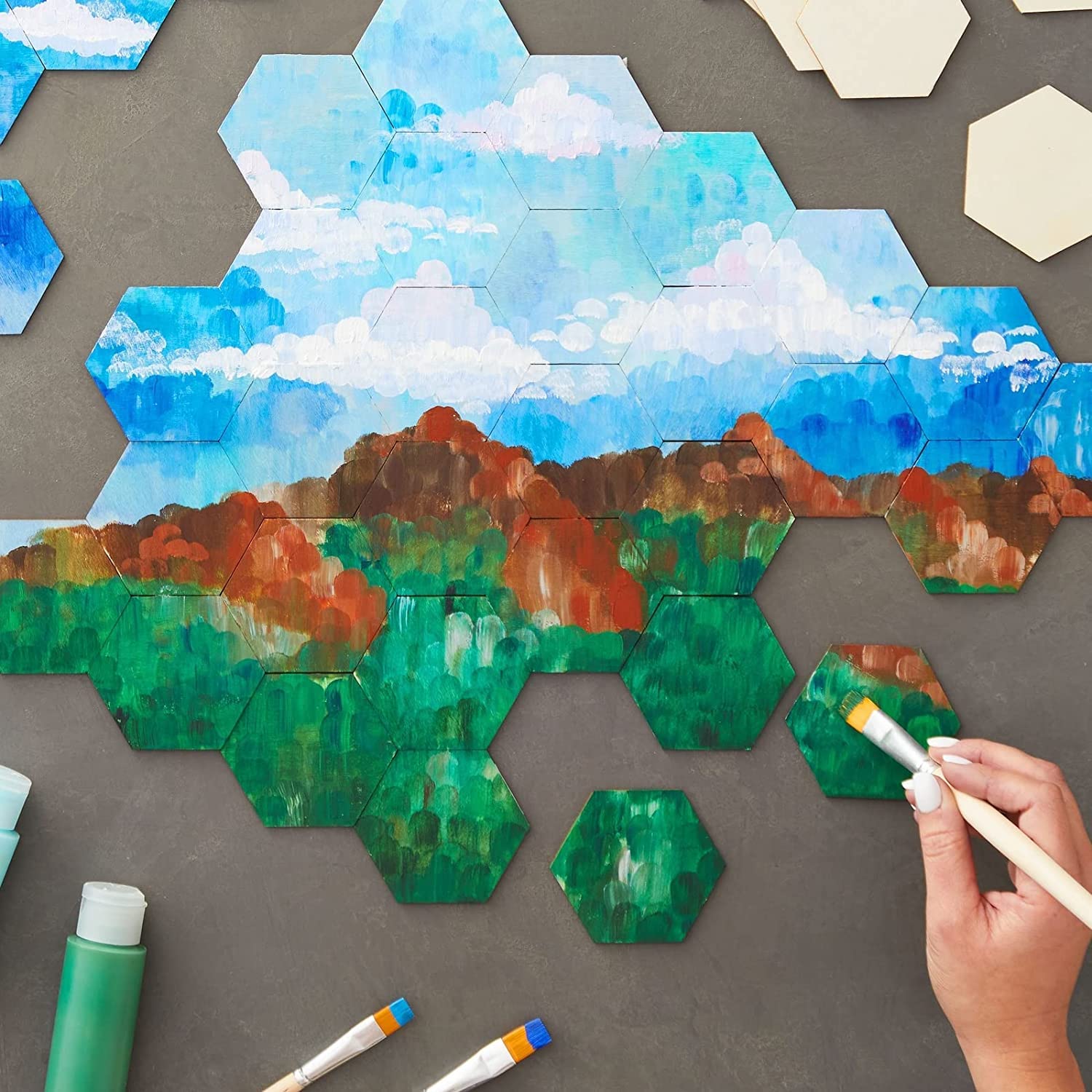 Hexagonal tiles with painted landscape scenes on a gray surface, with a hand holding a paintbrush.
