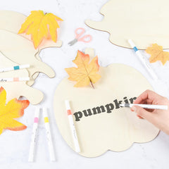 Handwriting 'pumpkin' on a leaf-shaped card with autumn leaves and markers on a white background