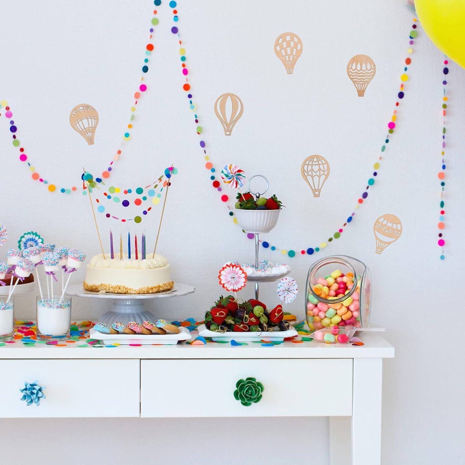Decorated birthday party table with cake, balloons, and colorful decorations against a white wall.