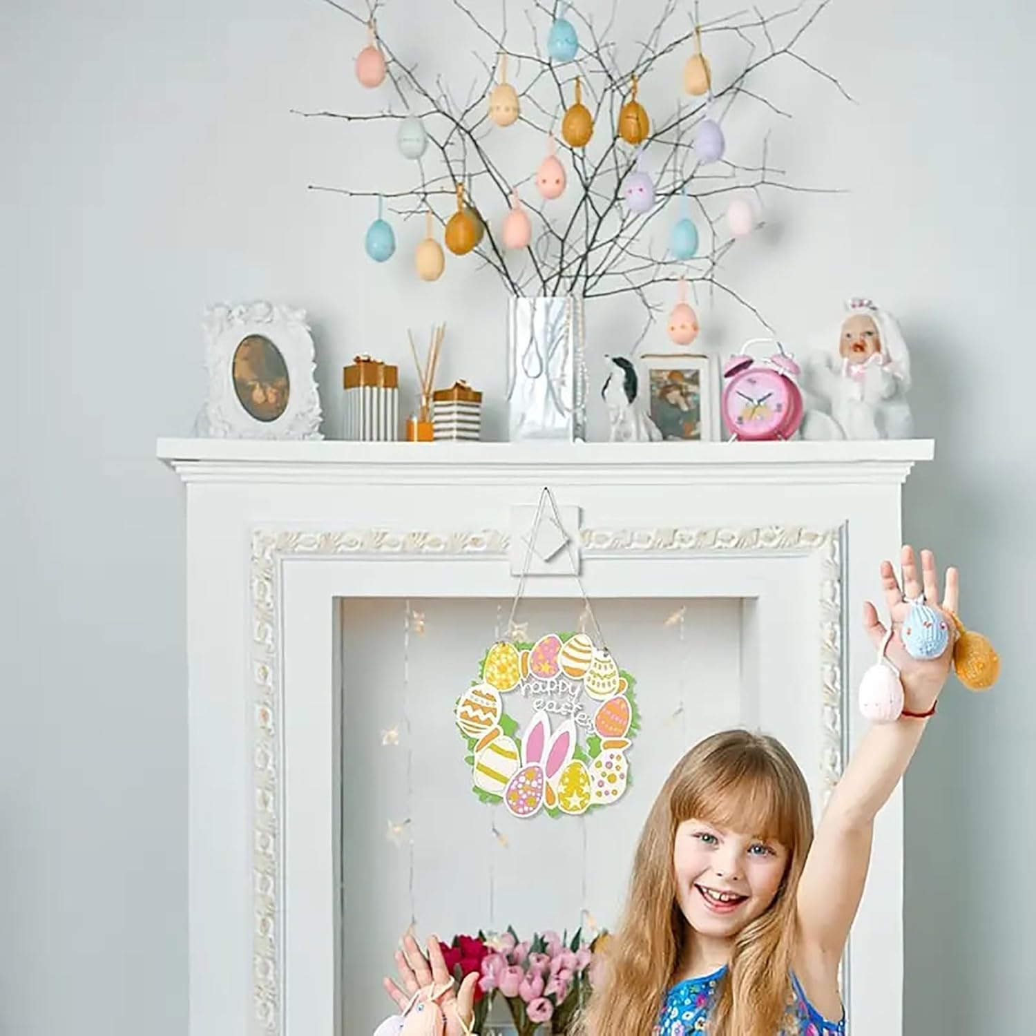 Child holding a small Easter decoration in front of a decorated fireplace with Easter eggs on branches.