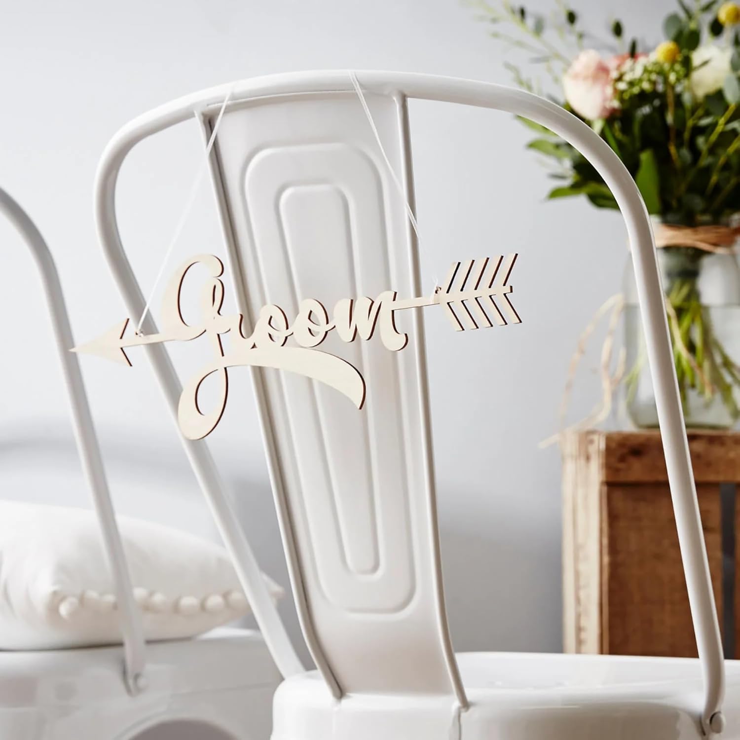 Wooden 'Groom' sign on a white chair with a blurred floral arrangement in the background
