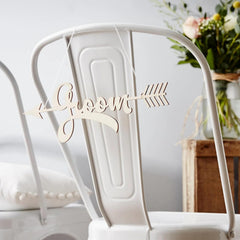 Wooden 'Groom' sign on a white chair with a blurred floral arrangement in the background