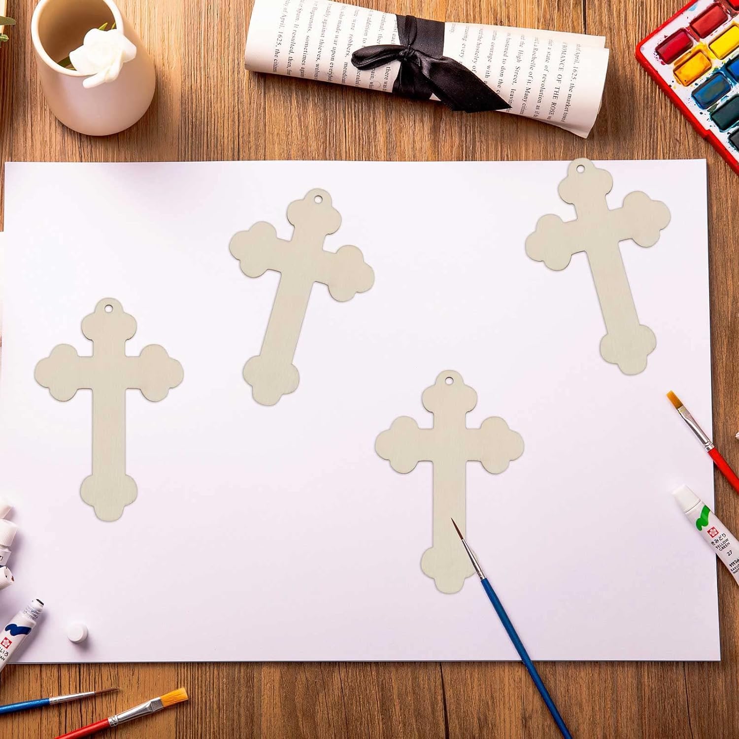 Cross-shaped wooden ornaments on a white sheet of paper with painting supplies on a wooden table.