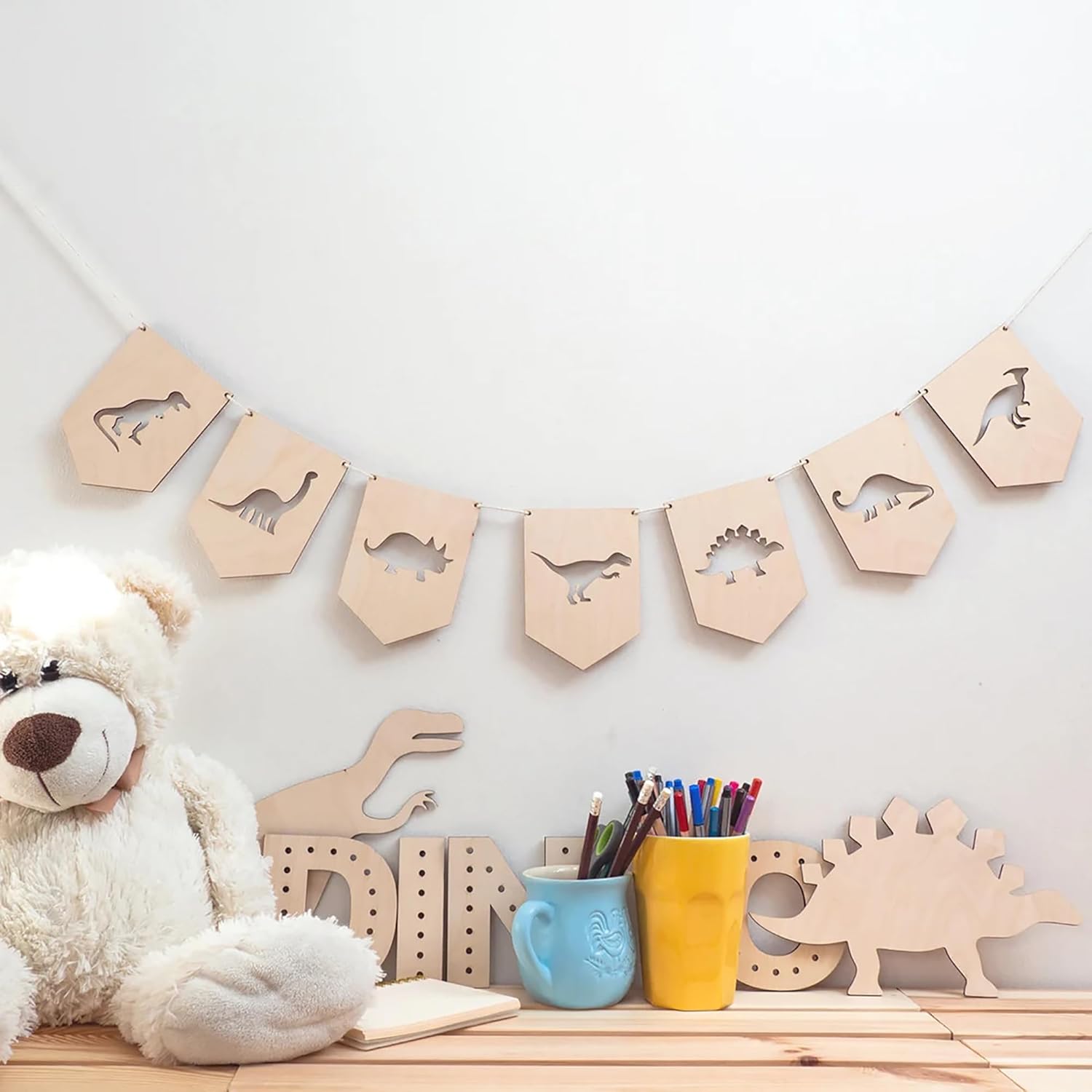 Wooden dinosaur banner on a wall with a teddy bear and stationery items below.