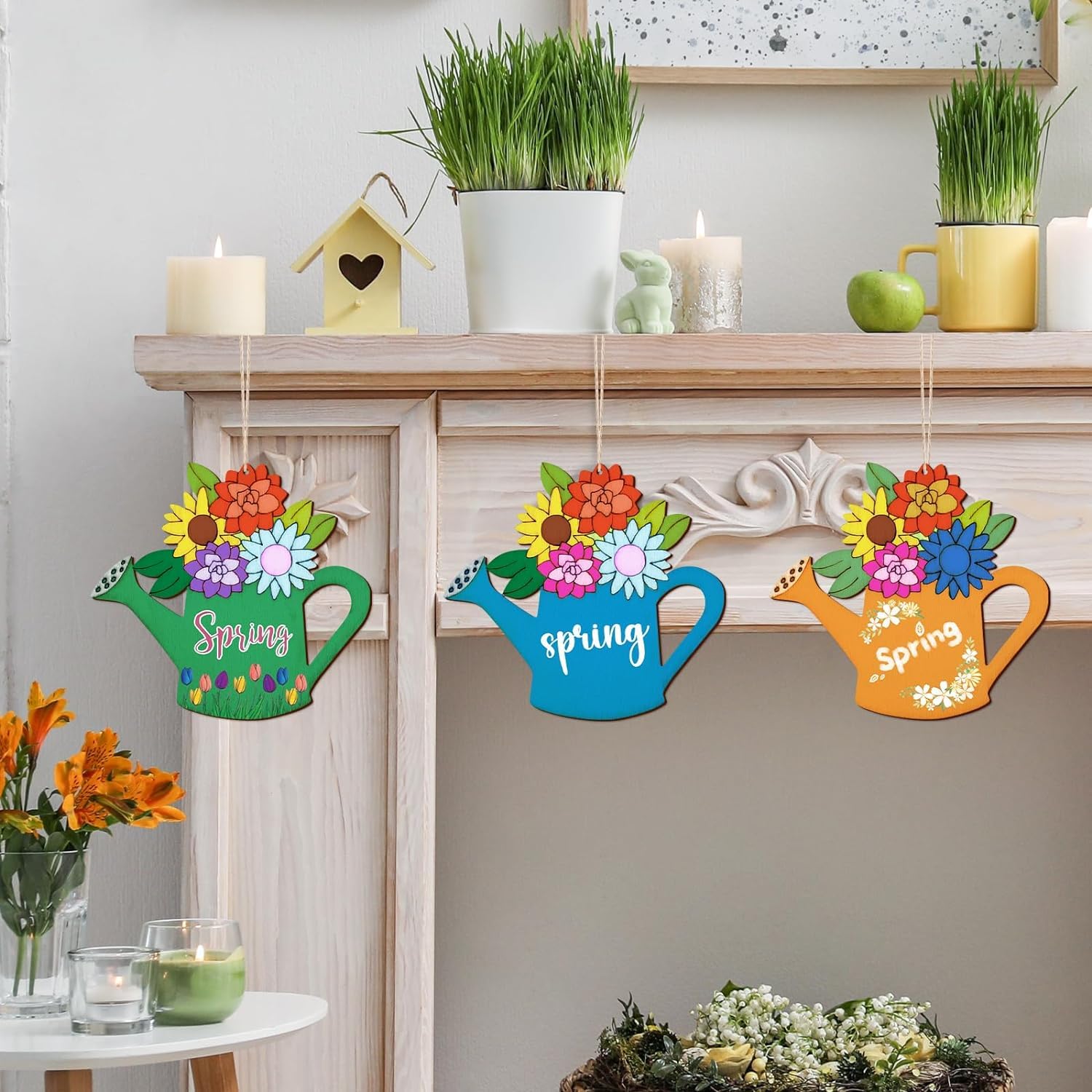 Colorful 'Spring' watering can decorations hanging on a wall with a fireplace and candles in the background.