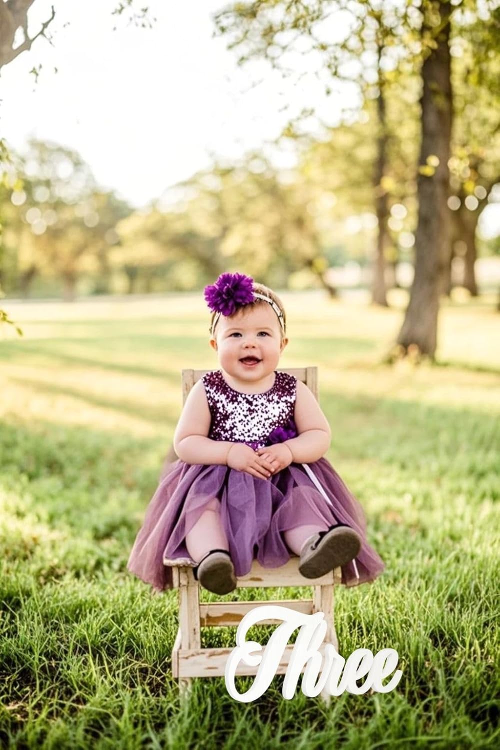 Child in a purple dress sitting on a chair with 'Three' sign in a park