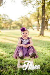 Child in a purple dress sitting on a chair with 'Three' sign in a park
