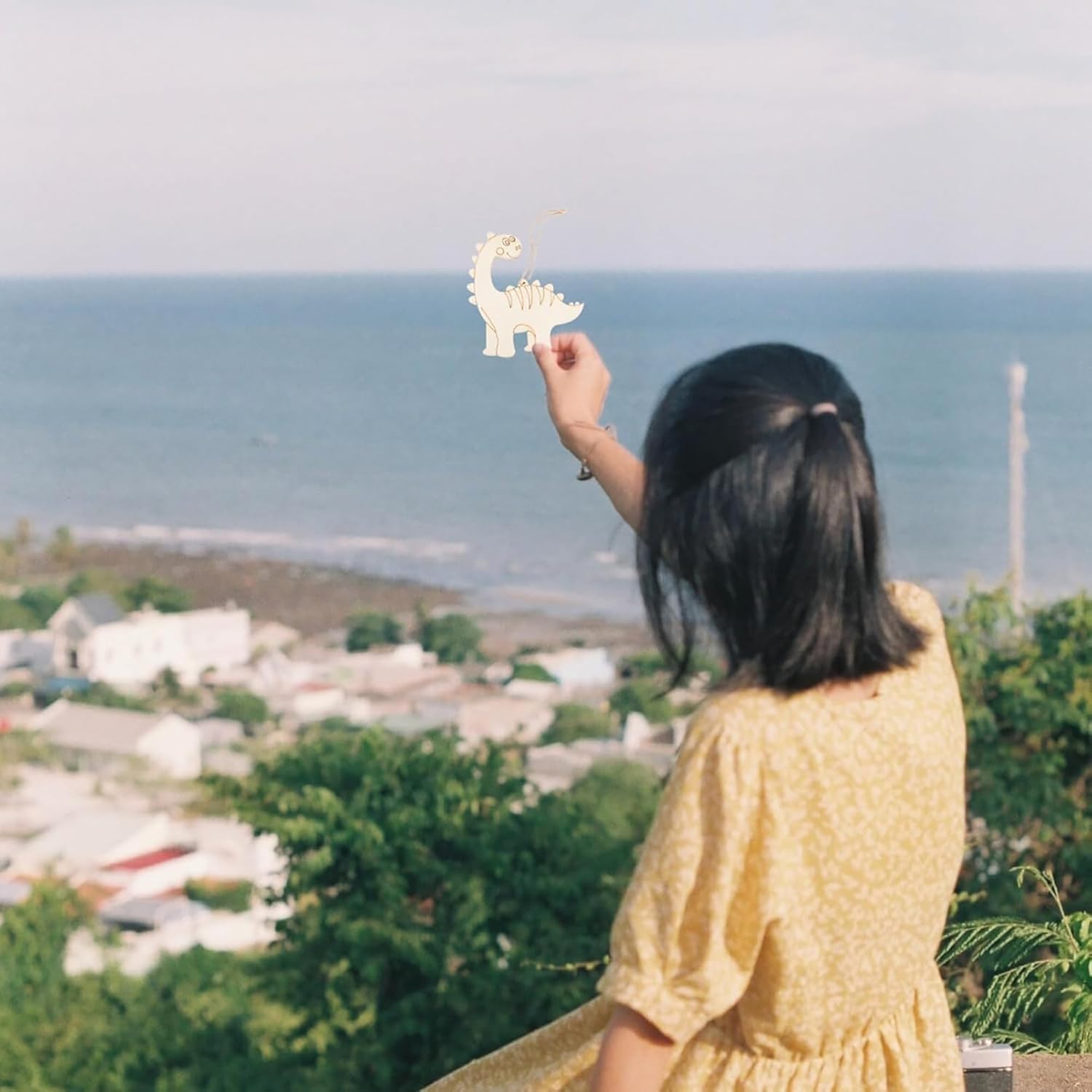 Woman in a yellow dress holding a dinosaur toy with a scenic view of the ocean and town in the background