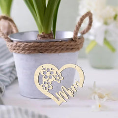 Heart-shaped decorative item with 'Mom' engraved, placed on a surface with a plant in the background.