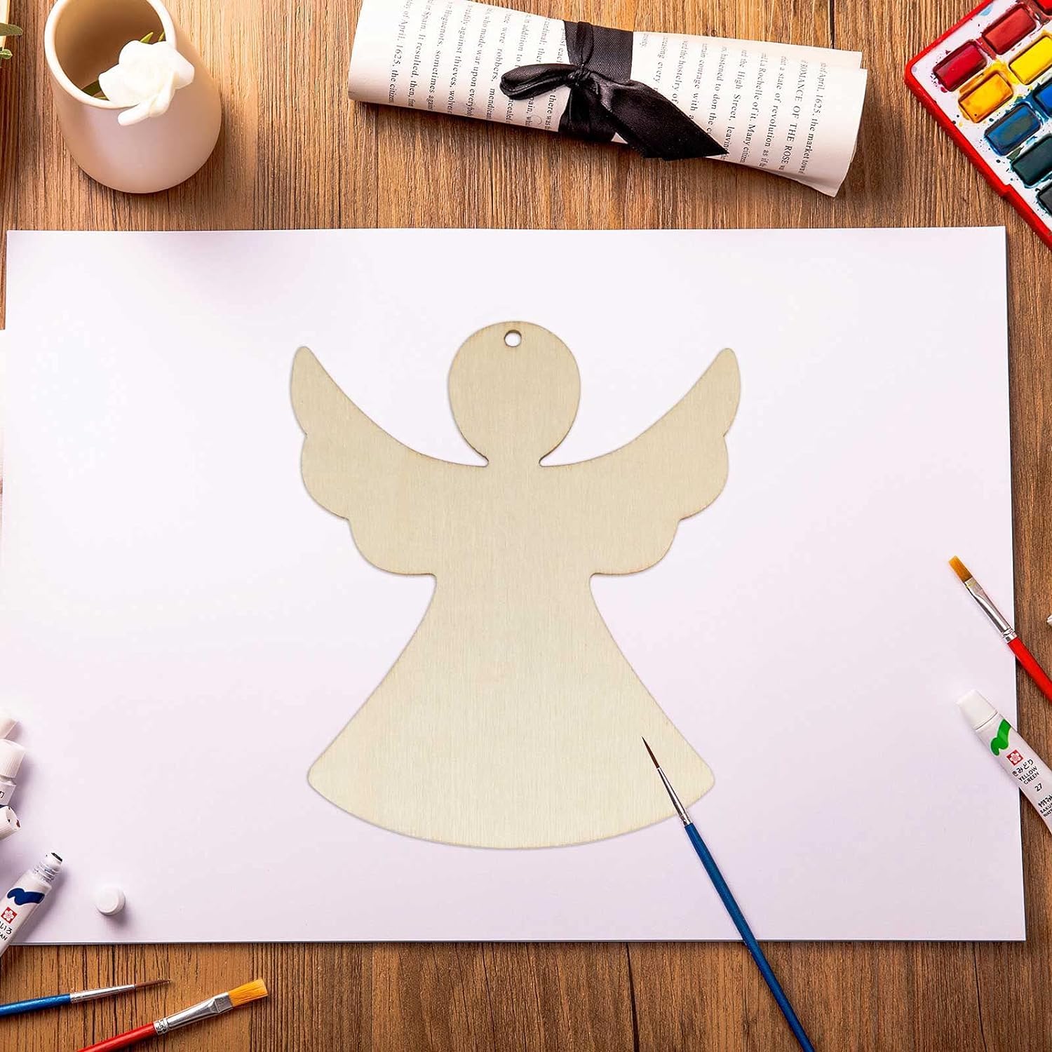 Angel-shaped wooden cutout on a white sheet of paper with art supplies on a wooden table.