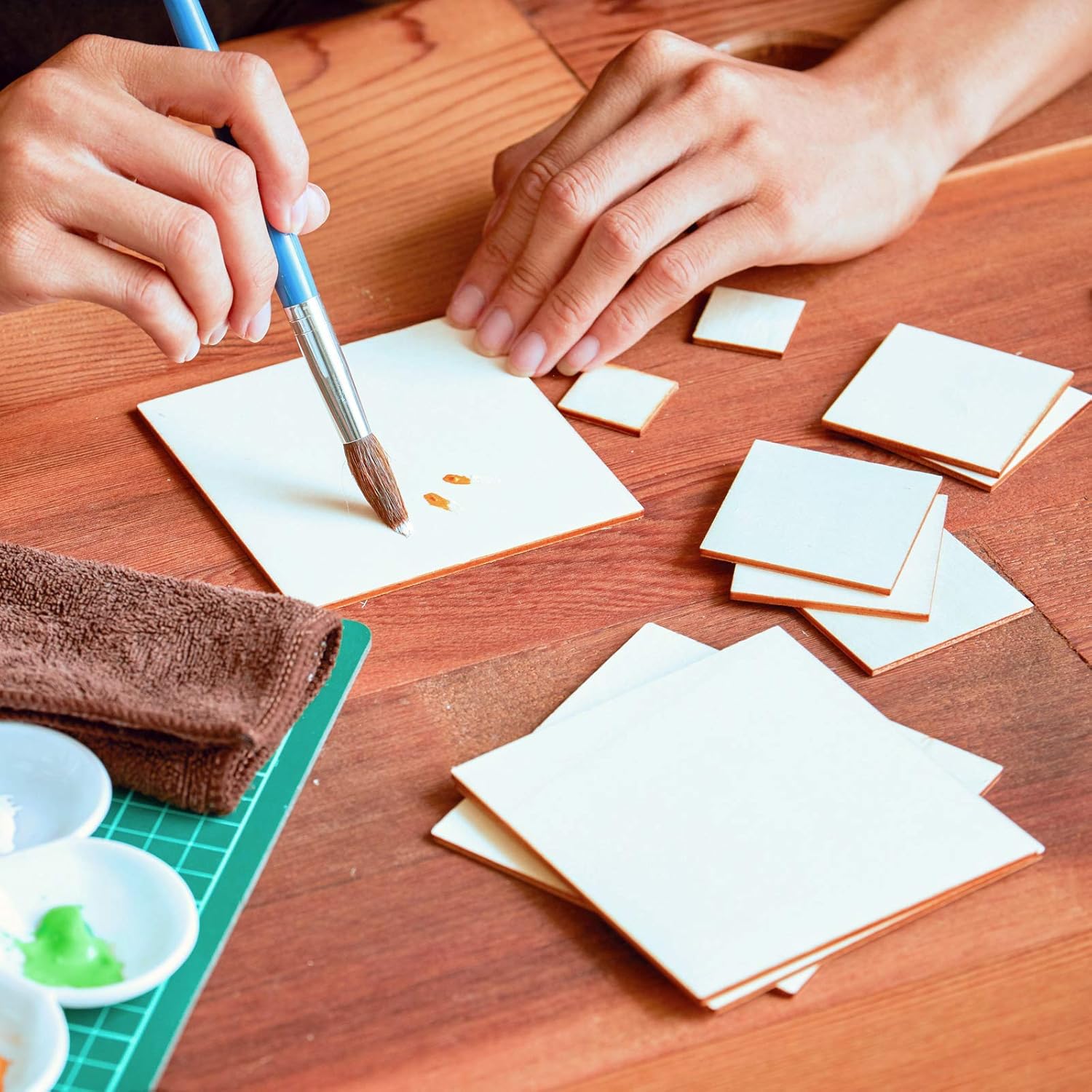 Hands using a brush to paint on small square pieces of paper on a wooden table.