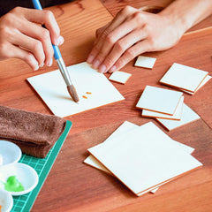 Hands using a brush to paint on small square pieces of paper on a wooden table.