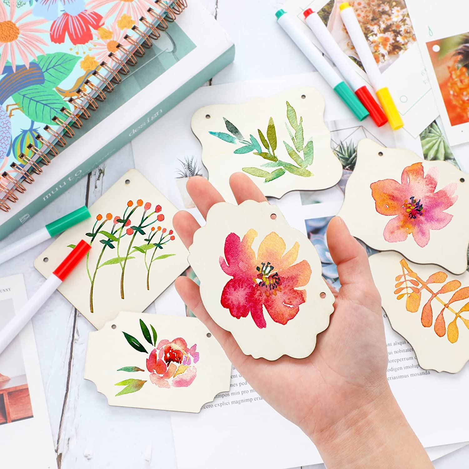 Hand holding a floral bookmark with watercolor designs on a desk with markers and notebooks.