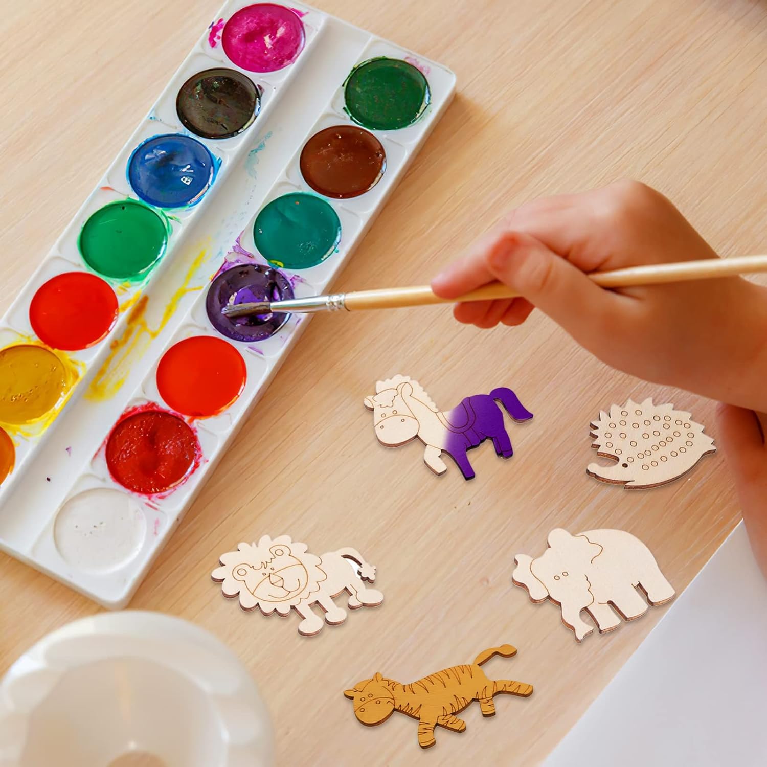 Hand painting wooden animal shapes with a watercolor palette on a wooden table.