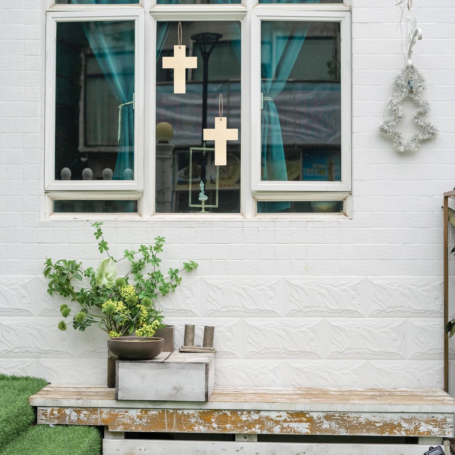 Decorative outdoor setting with plants, crosses, and a bench against a white wall.