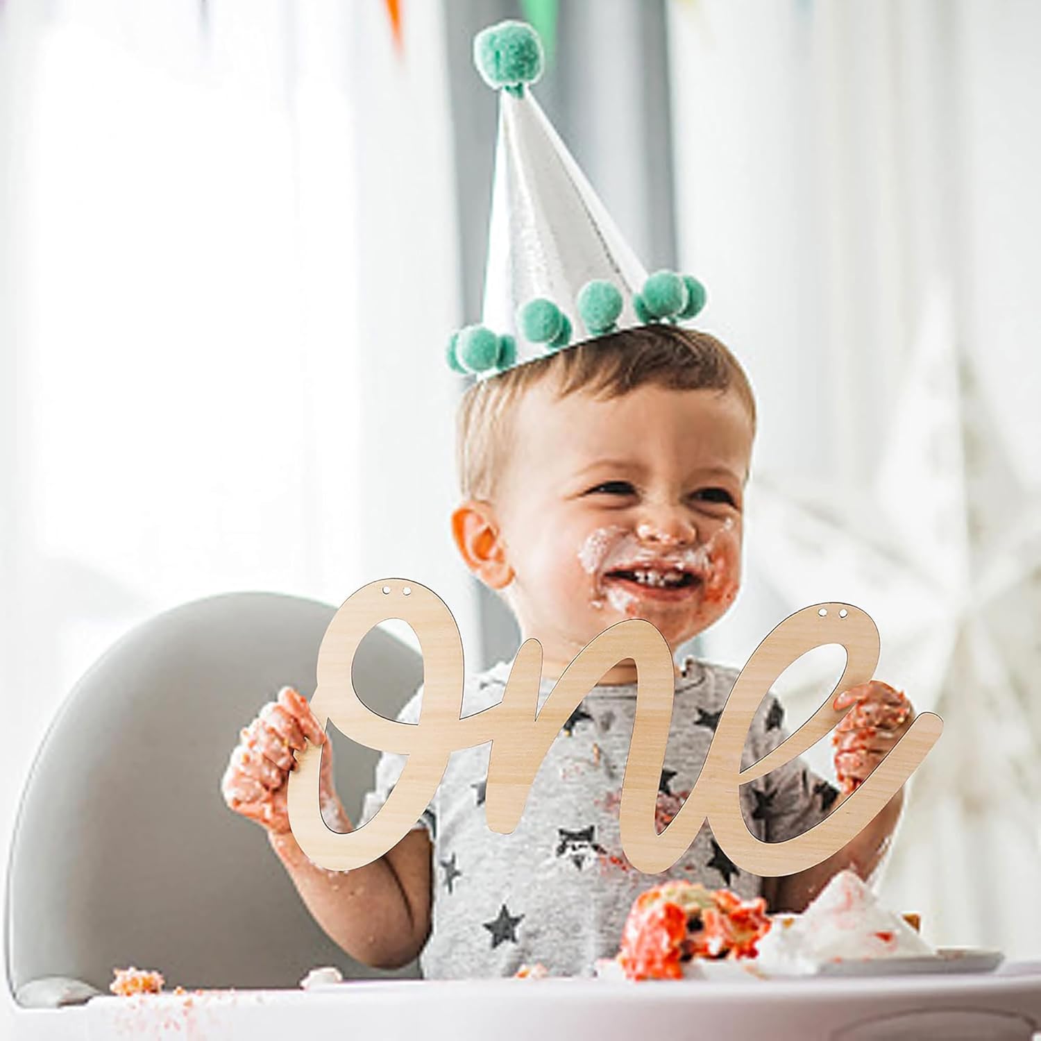 Child celebrating a first birthday with a party hat and 'one' sign, surrounded by cake and a high chair.