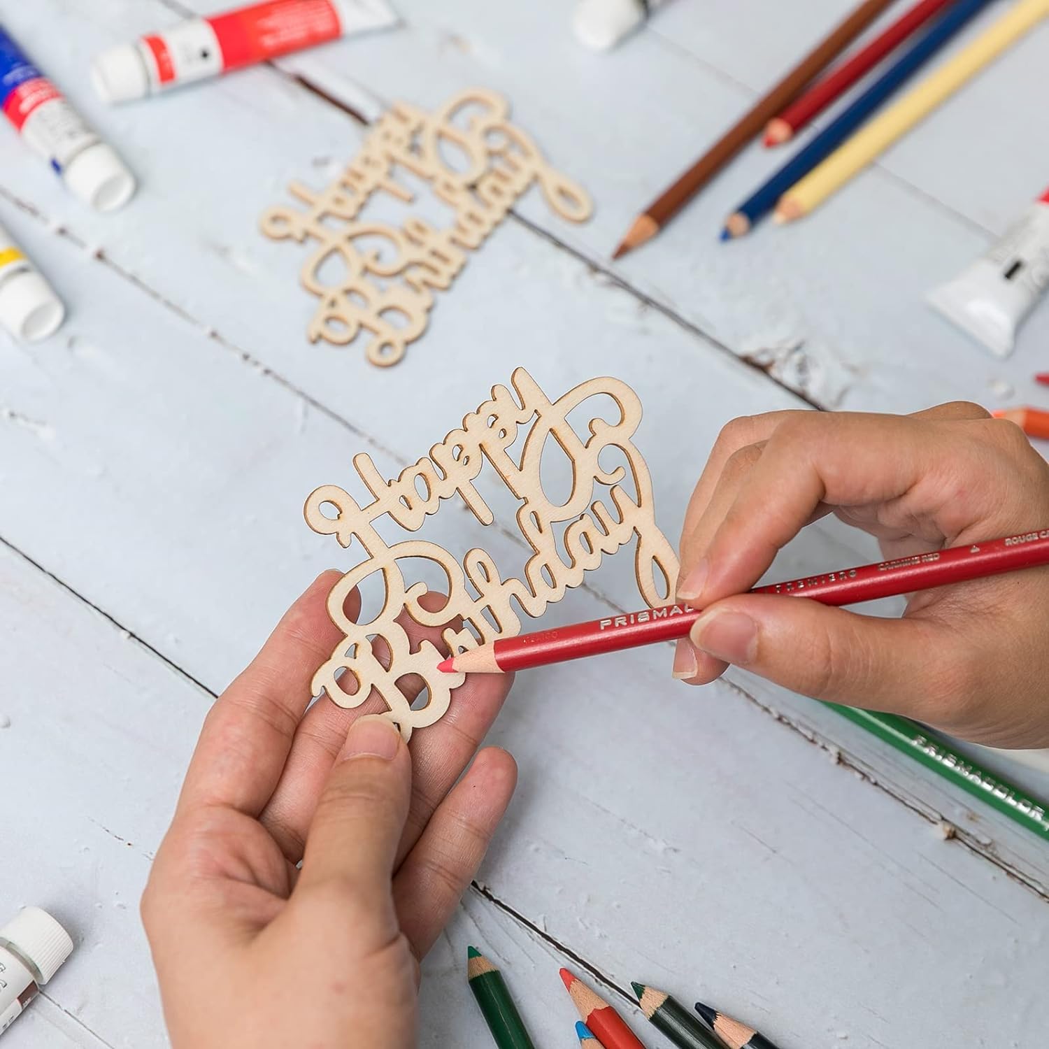 Person holding a red pencil next to a wooden 'Happy Birthday' decoration on a light blue surface with stationery items.