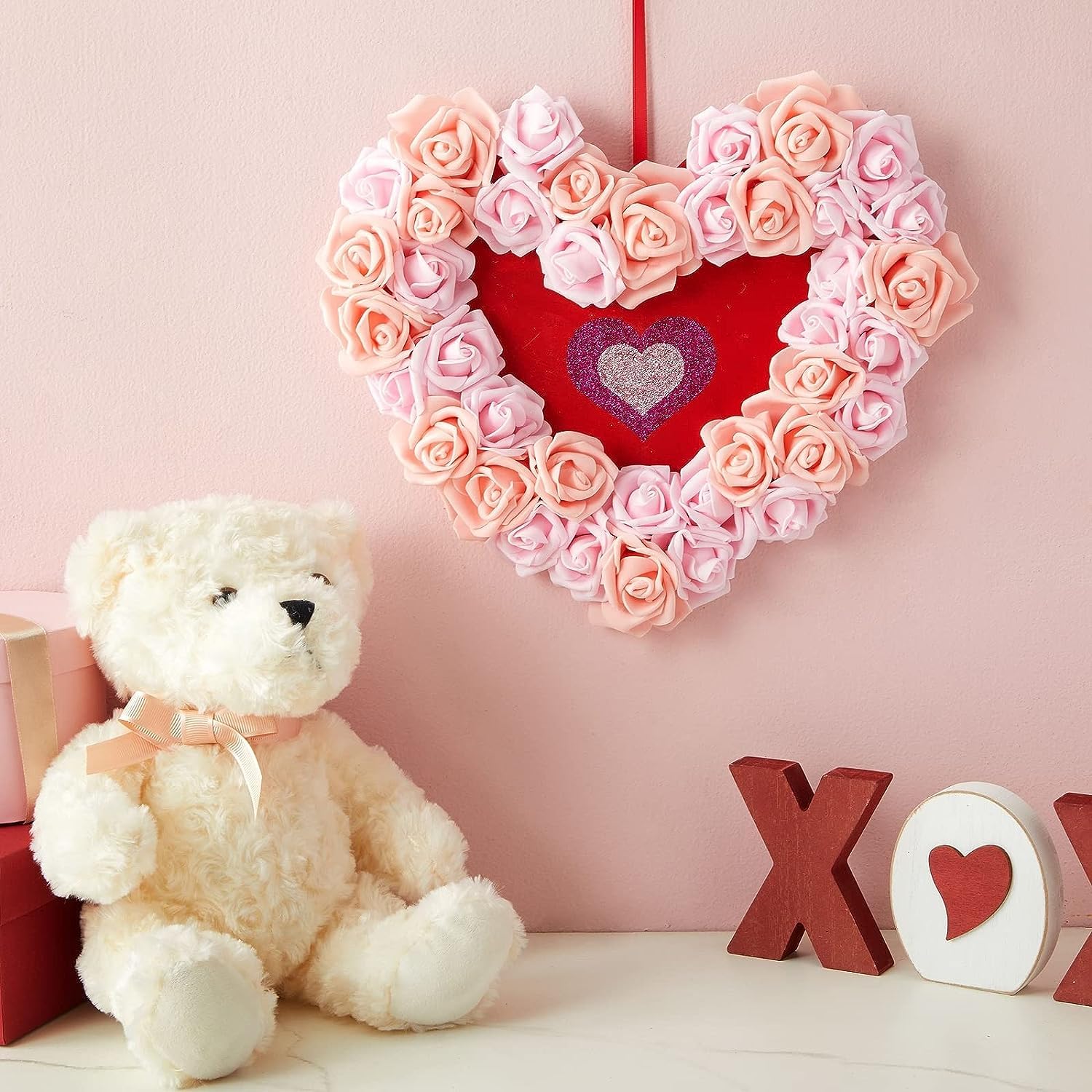 Heart-shaped arrangement of pink and red roses with a teddy bear and decorative letters on a pink background