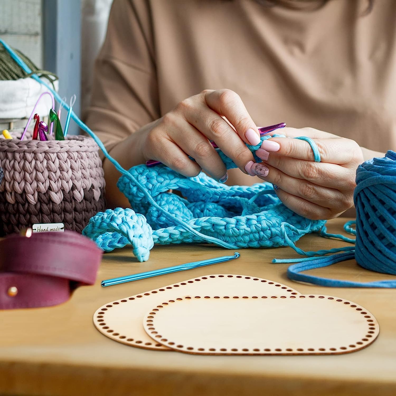 Person knitting blue yarn on a wooden table with a basket of yarn and a pattern book.