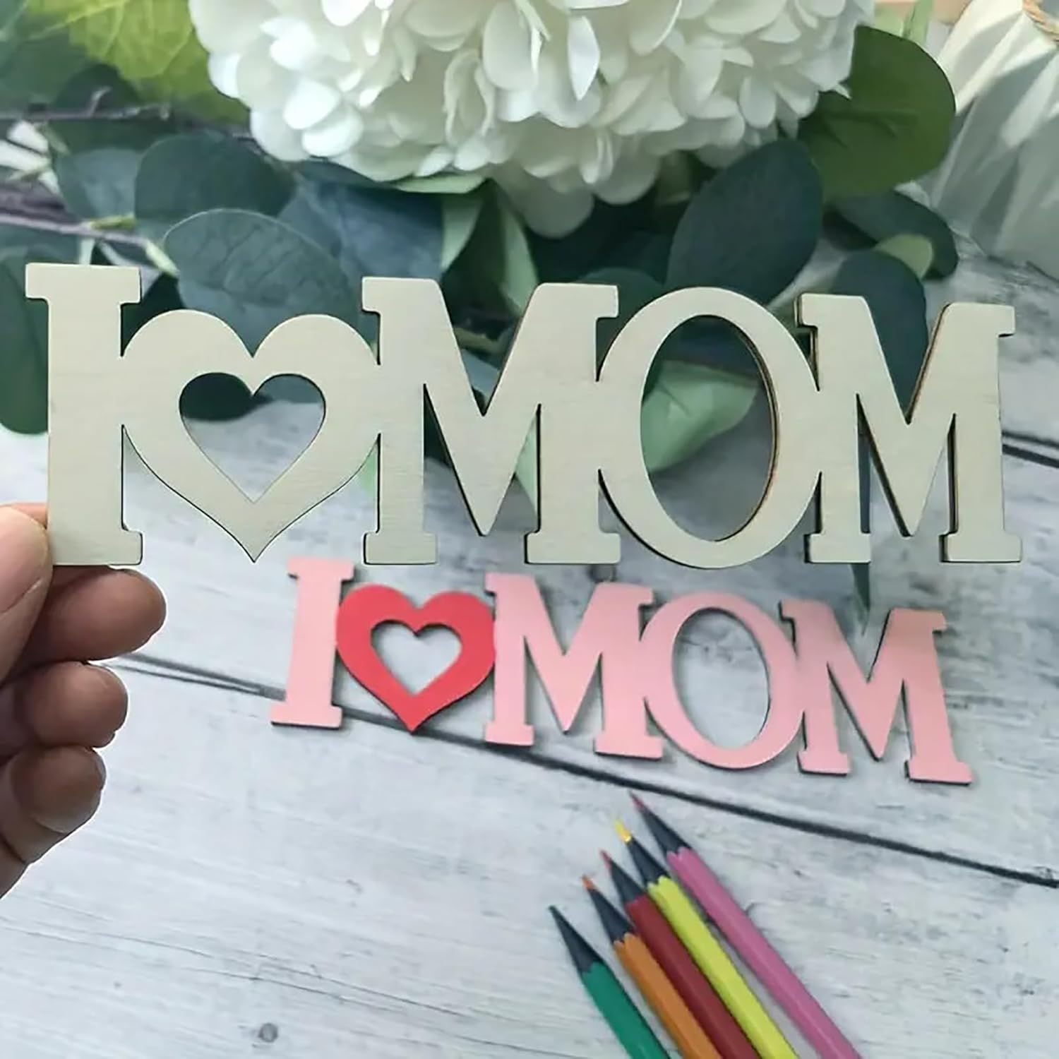 Wooden 'I ❤️ MOM' sign held by a hand with flowers and colored pencils in the background.