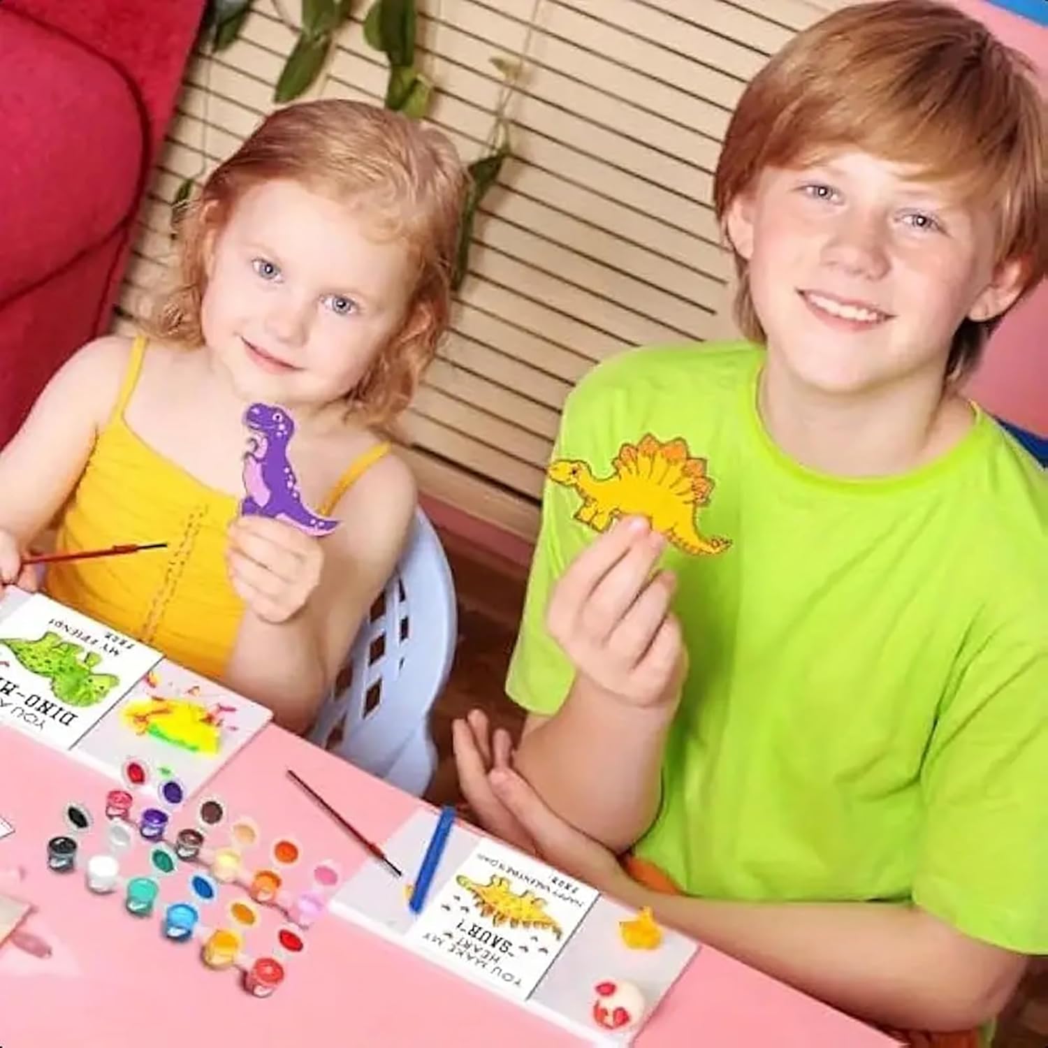 Two children sitting at a table with art supplies, including paint and cards with dinosaur designs.