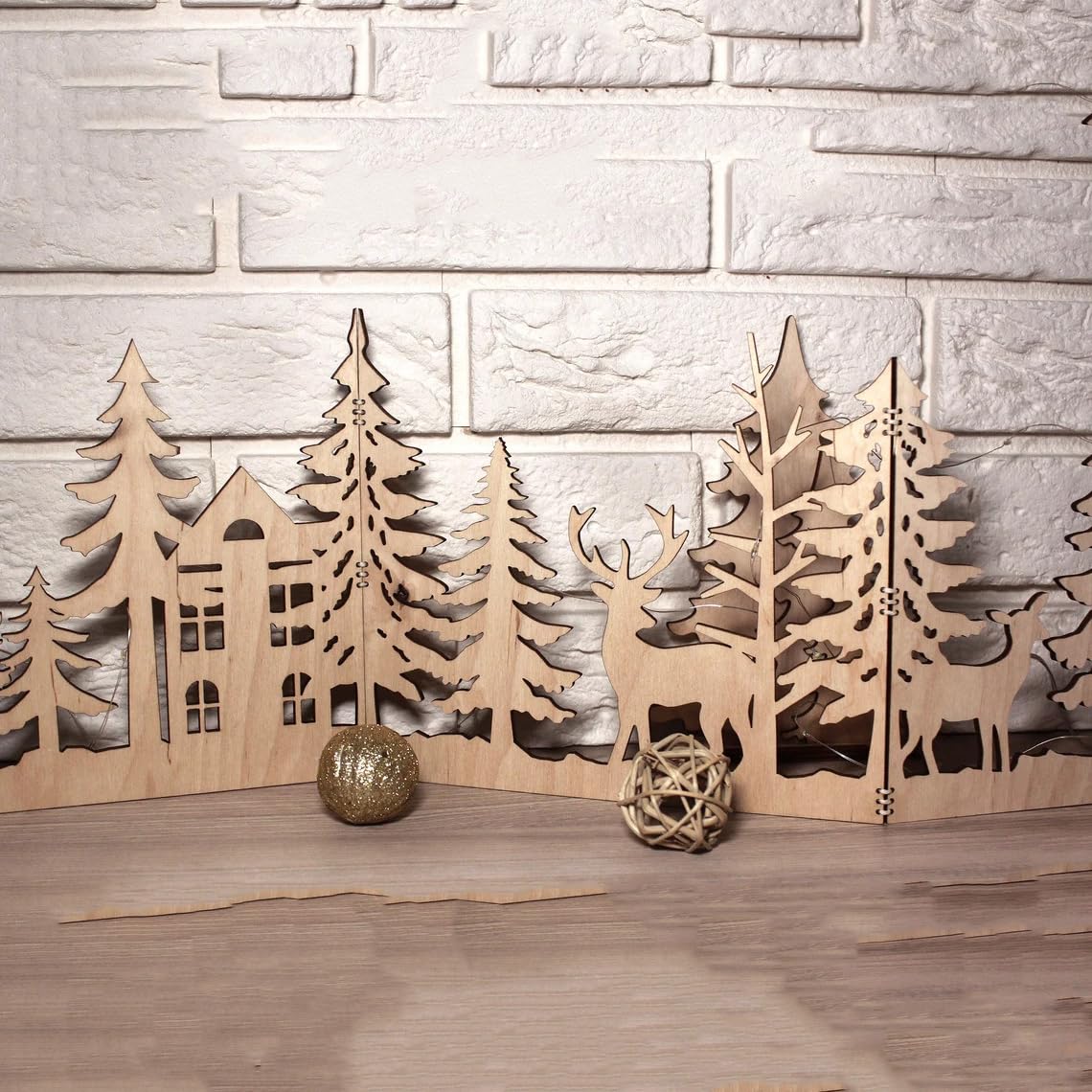 Wooden cutout decorations of trees, houses, and deer on a wooden surface with a stone wall background.