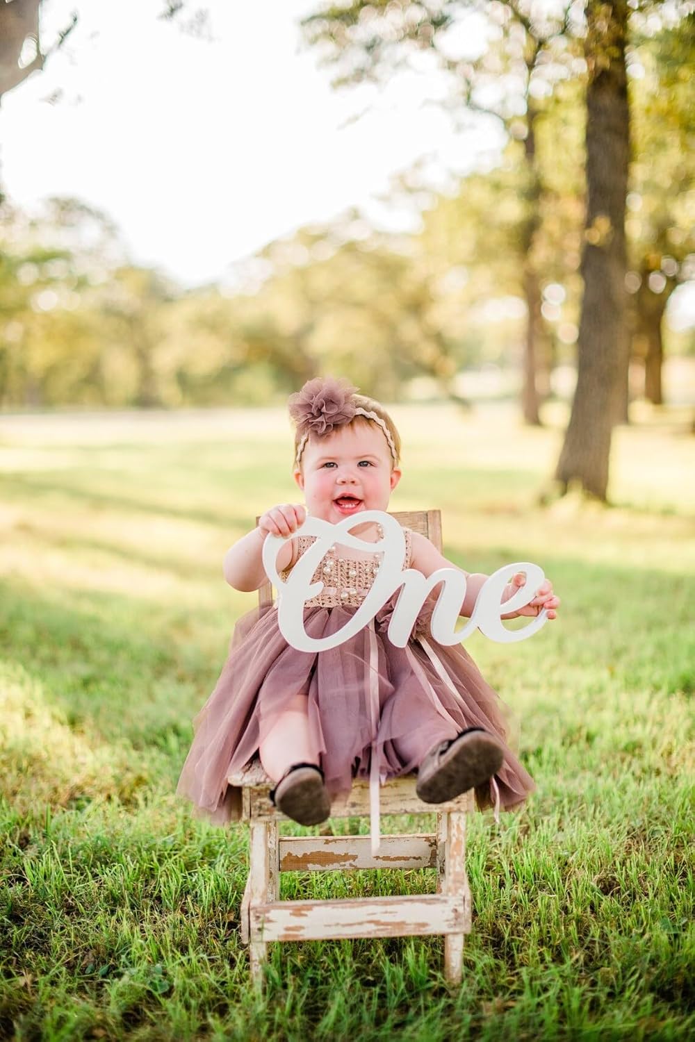 Child in a pink dress sitting on a small stool with a 'One' sign outdoors.
