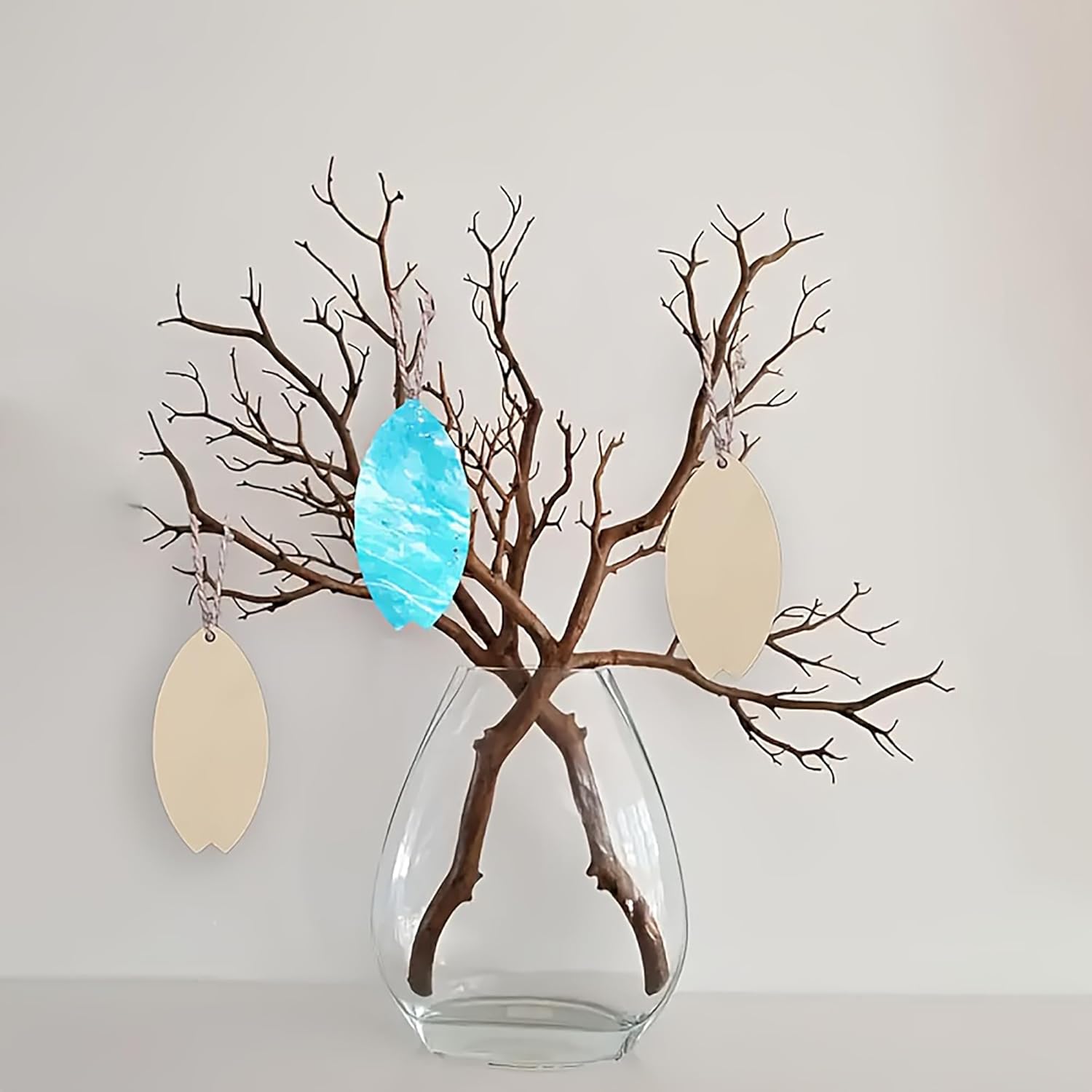 Decorative arrangement with branches, a blue ornament, and beige leaves in a clear vase on a light gray background.