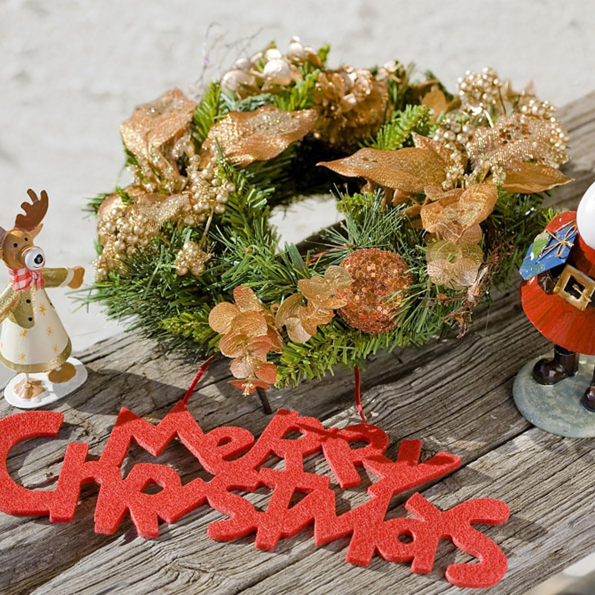 Decorative Christmas wreath with gold accents on a wooden surface, accompanied by festive figurines and 'Merry Christmas' sign.