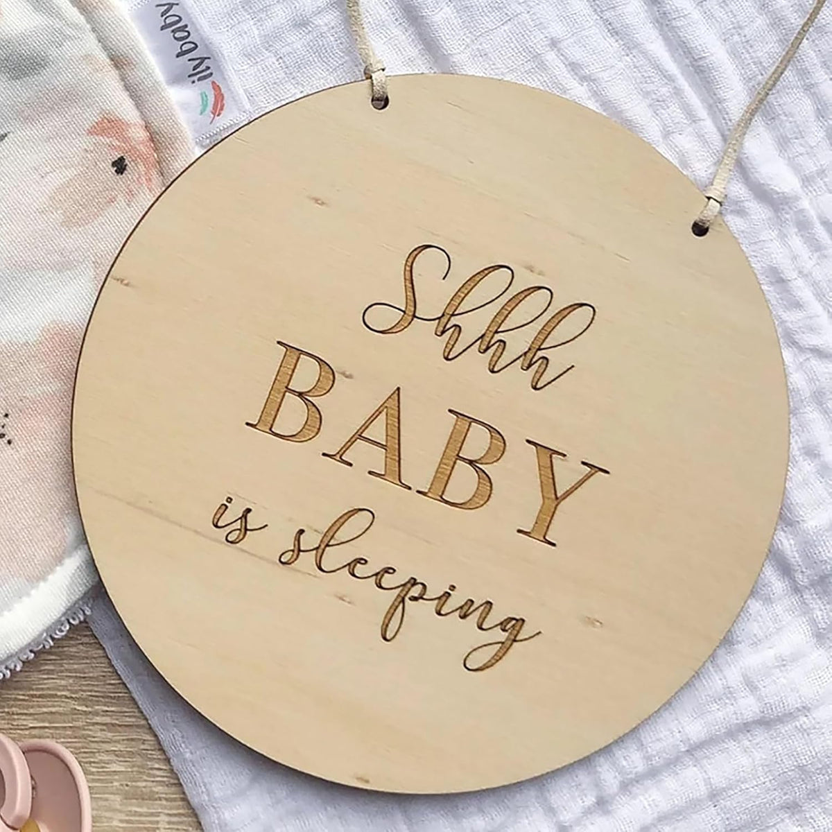 Round wooden sign with 'Shhh BABY is sleeping' text on a textured fabric background