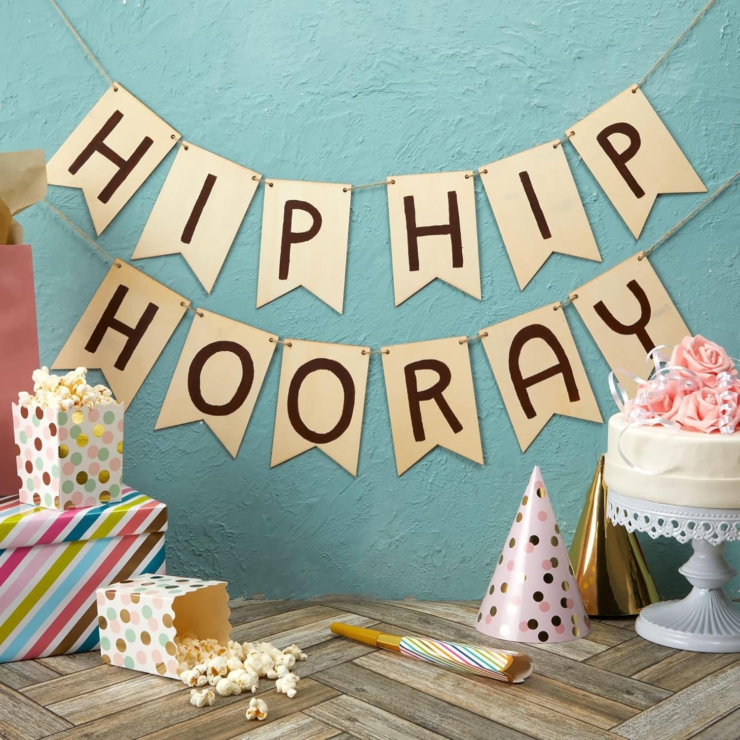 Party setup with 'HIP HIP HOORAY' banner, gifts, cake, and popcorn on a wooden floor against a blue wall.