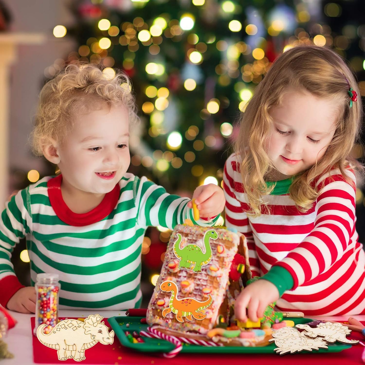 Two children in striped shirts decorating gingerbread houses with a Christmas tree in the background.
