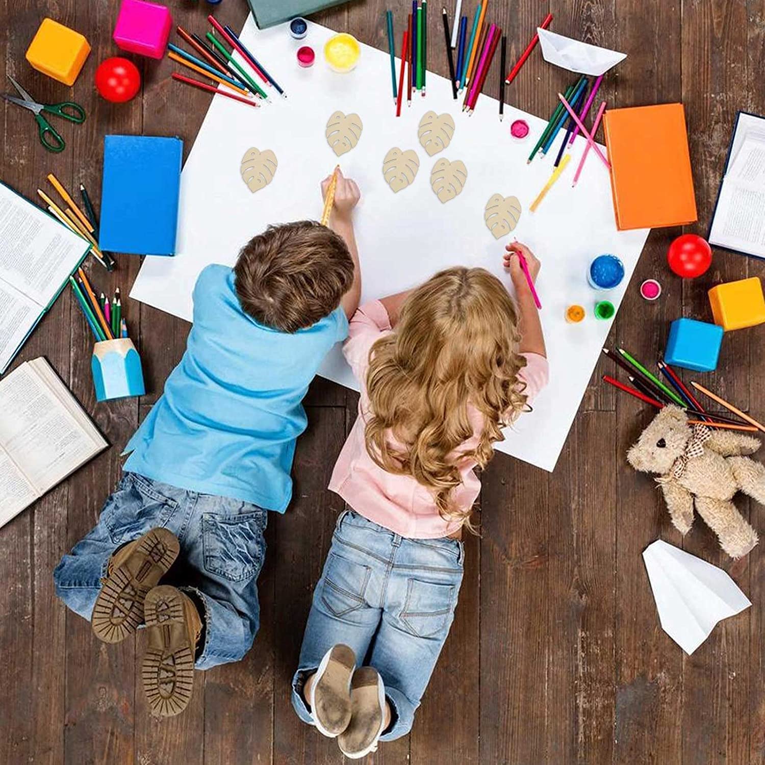 Two children drawing on a large sheet of paper with various art supplies on a wooden floor.