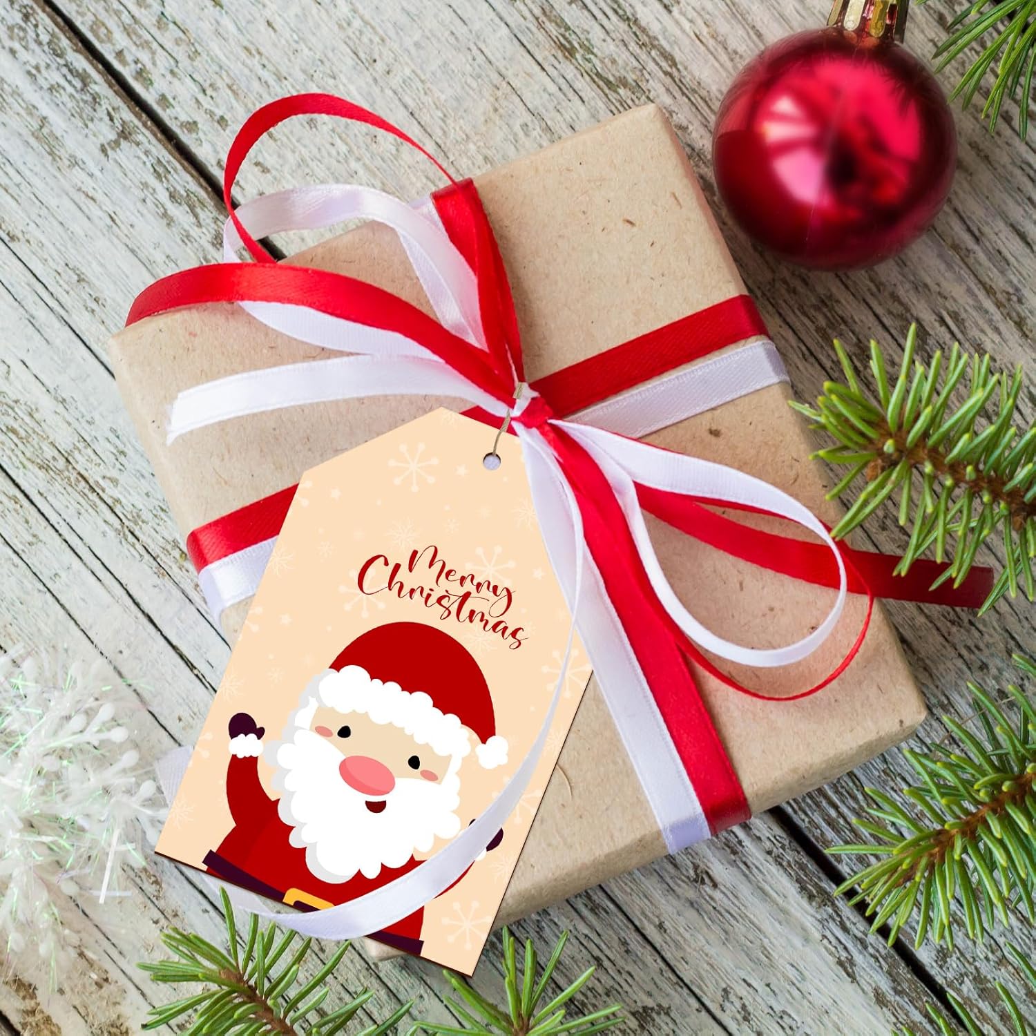 Gift wrapped in brown paper with a red ribbon and Santa Claus tag on a wooden surface with Christmas decorations.