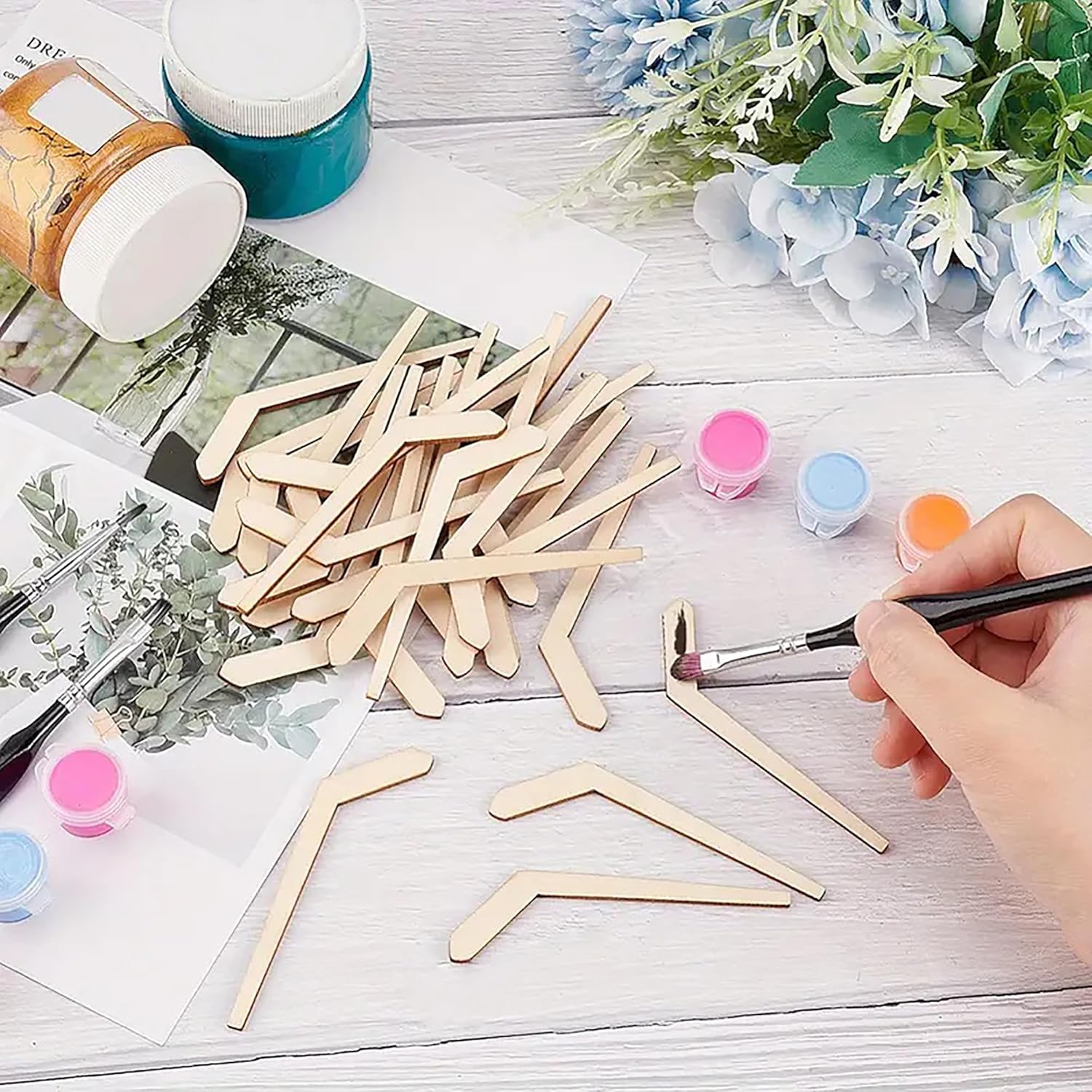 Person painting wooden sticks with a brush on a table with paint containers and floral decorations.