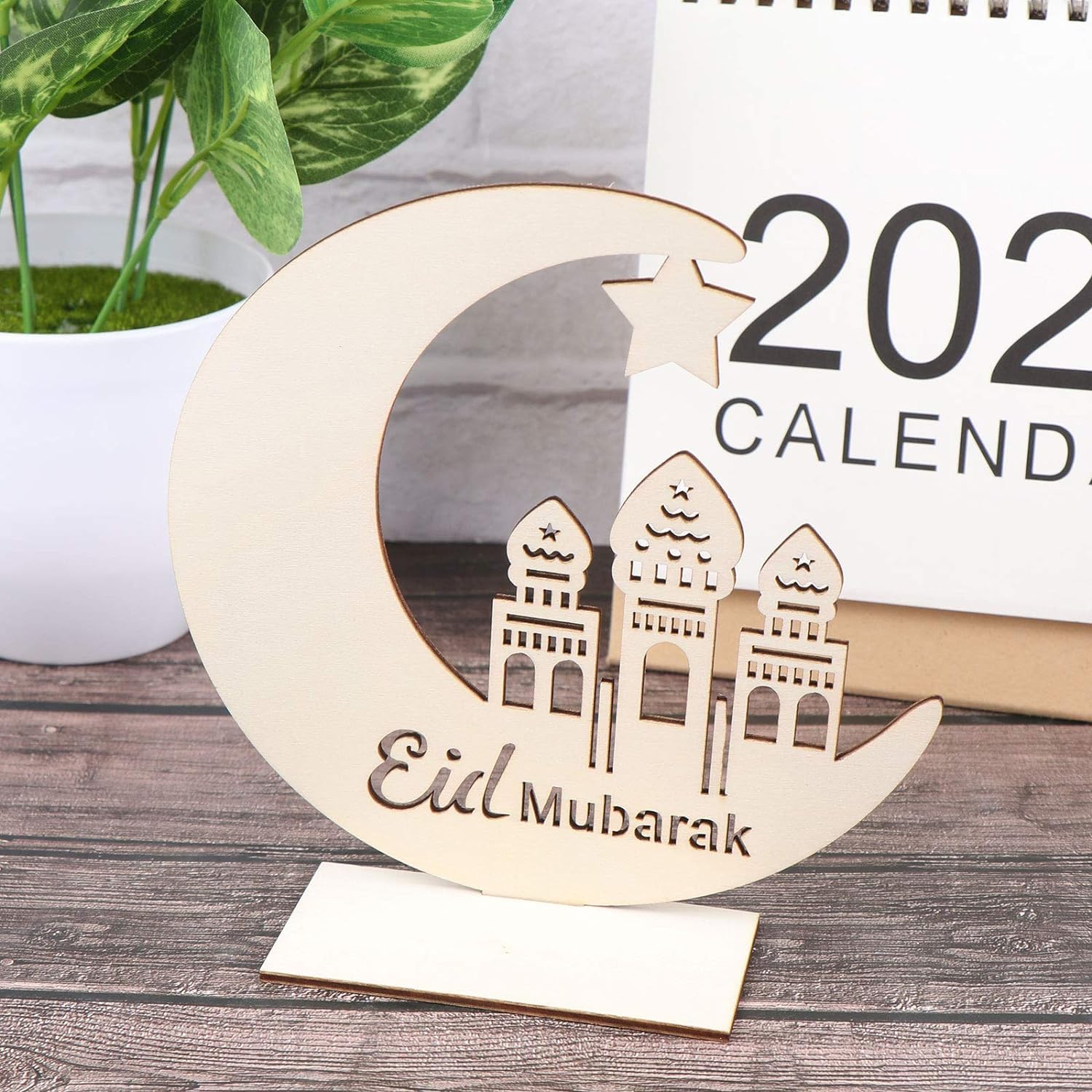 Wooden crescent moon with 'Eid Mubarak' and mosque silhouette on a wooden surface with a calendar in the background.