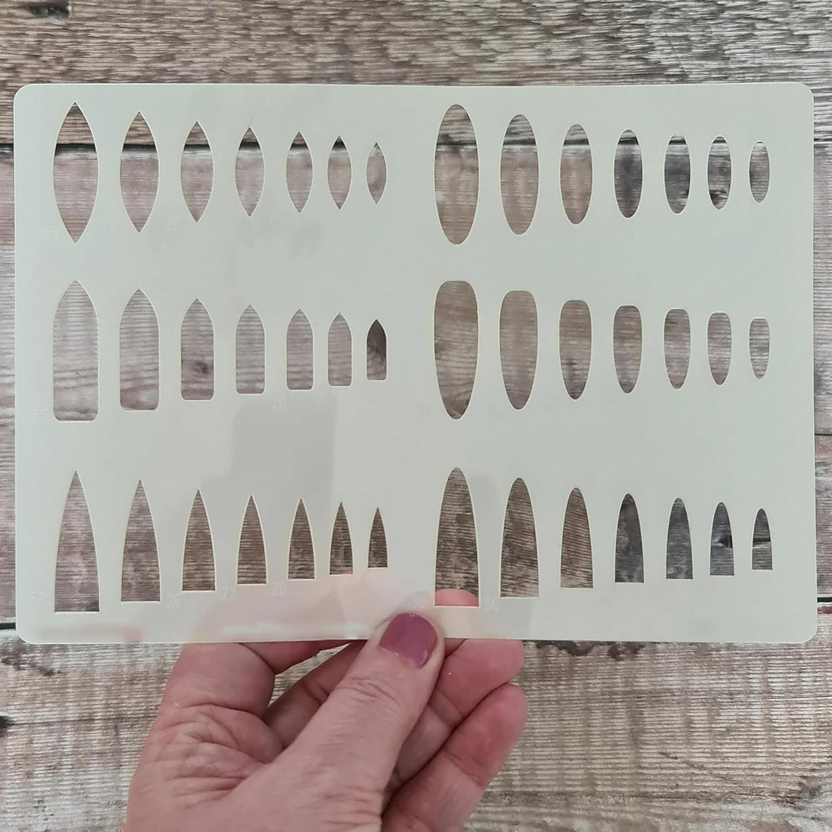 Hand holding a white stenciling template with geometric cutouts on a wooden surface