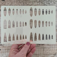 Hand holding a white stenciling template with geometric cutouts on a wooden surface