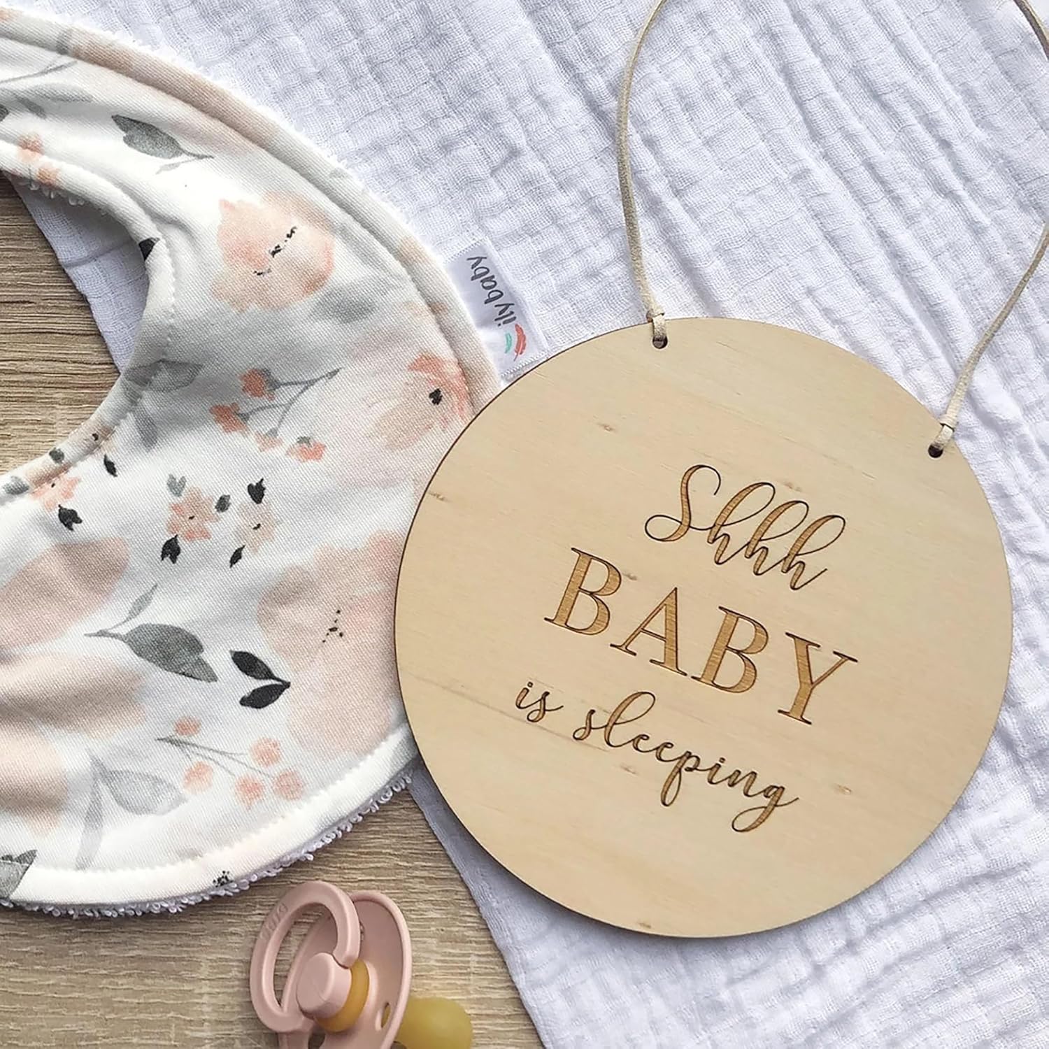 Wooden sign with 'Shhh BABY is sleeping' text next to a floral baby blanket and pink pacifier.