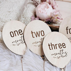 Wooden circular markers labeled 'one month', 'two months', and 'three months' on a soft surface with flowers.