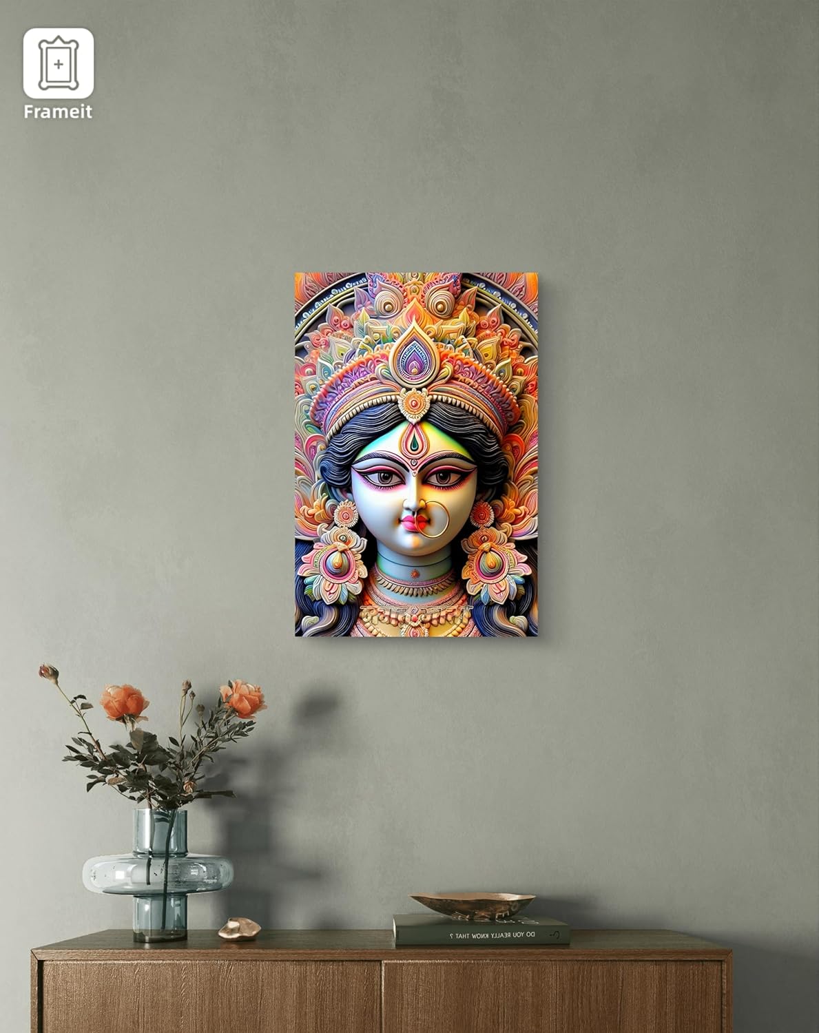 Decorative wall art of a colorful deity head on a gray wall above a wooden cabinet with a vase and books.