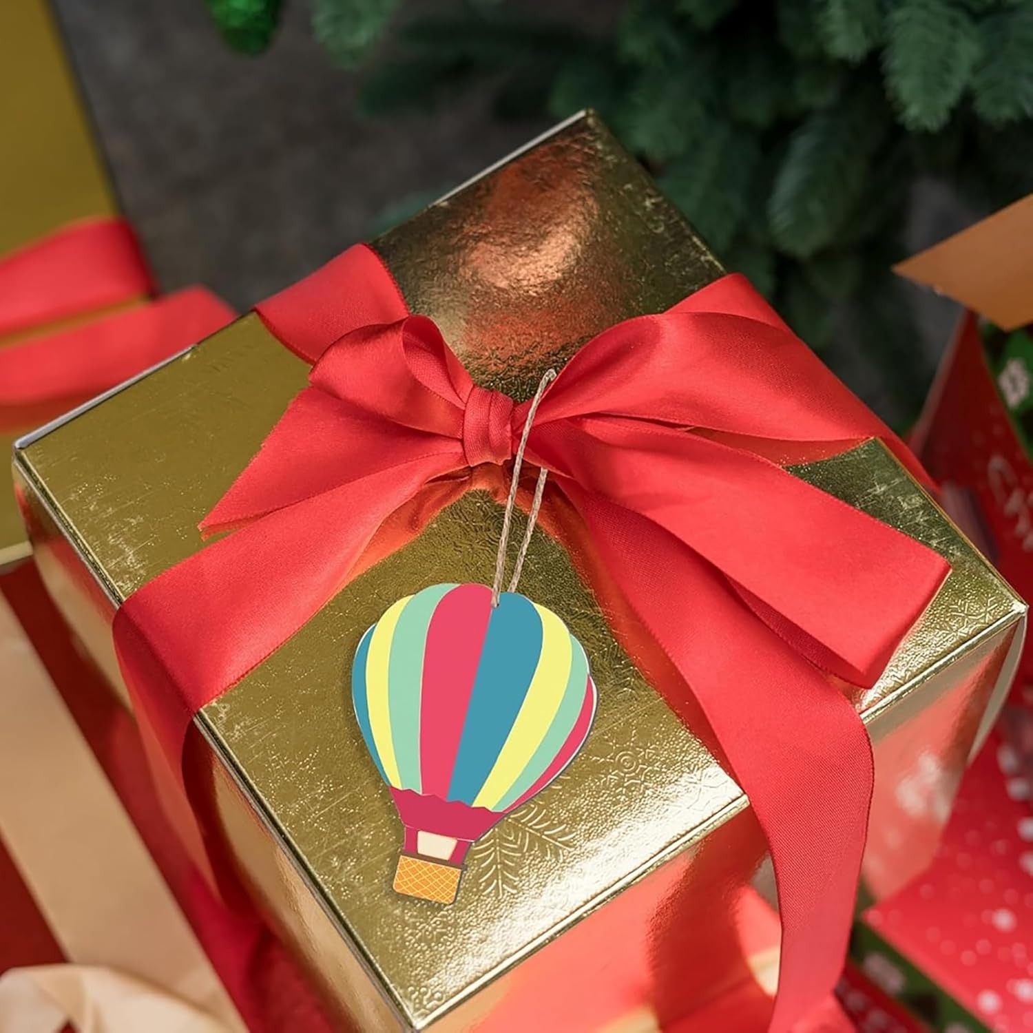 Gold gift box with a colorful hot air balloon ornament and red ribbon, set against a blurred festive background.